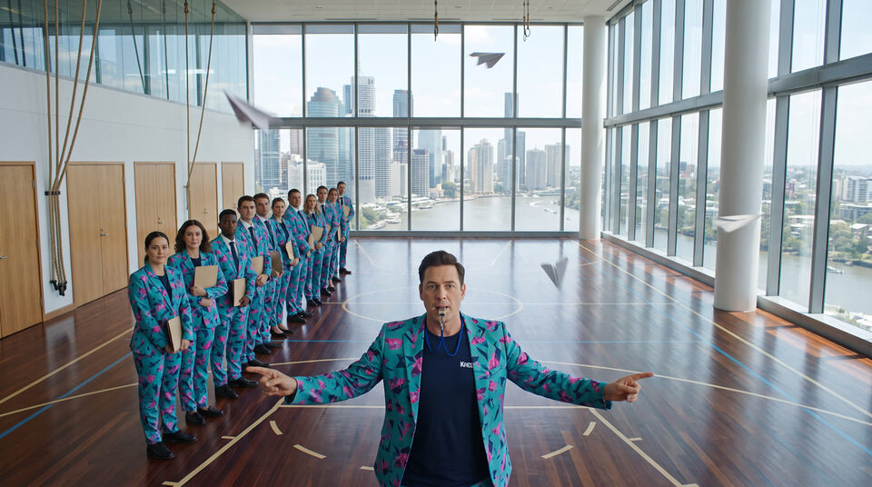 An instructor in teal print in an empty hall, with a line of trainees in matching suits behind him