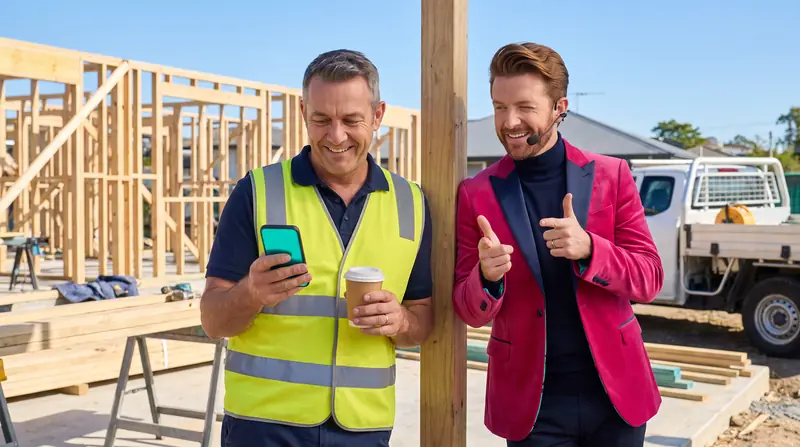 The owner grins at his phone on a building site while Monty, the AI coach, leans on a post behind him with finger guns.