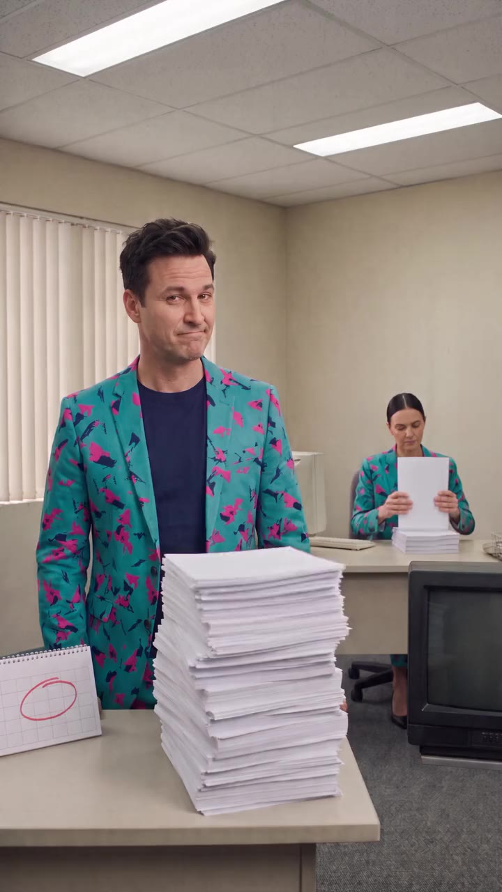 The first frame of the film: Buddy (AI) stands behind a tall stack of paper on an office desk while an AI staffer reads a page at the back of the room.