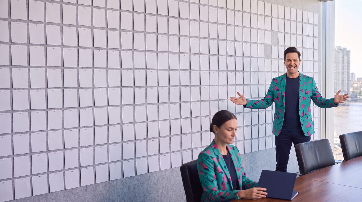 Buddy (AI) stands arms wide at a boardroom wall covered in blank cards while an AI staffer works at a laptop, with the Brisbane River through the glass.