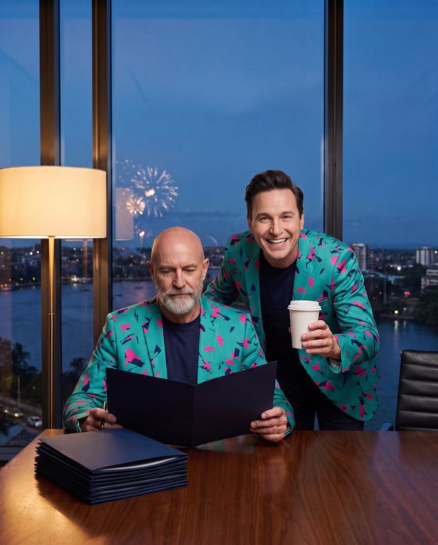 An AI staffer reads a proposal in a navy folder at an evening desk by the river while Buddy (AI) stands behind him with a coffee, grinning.
