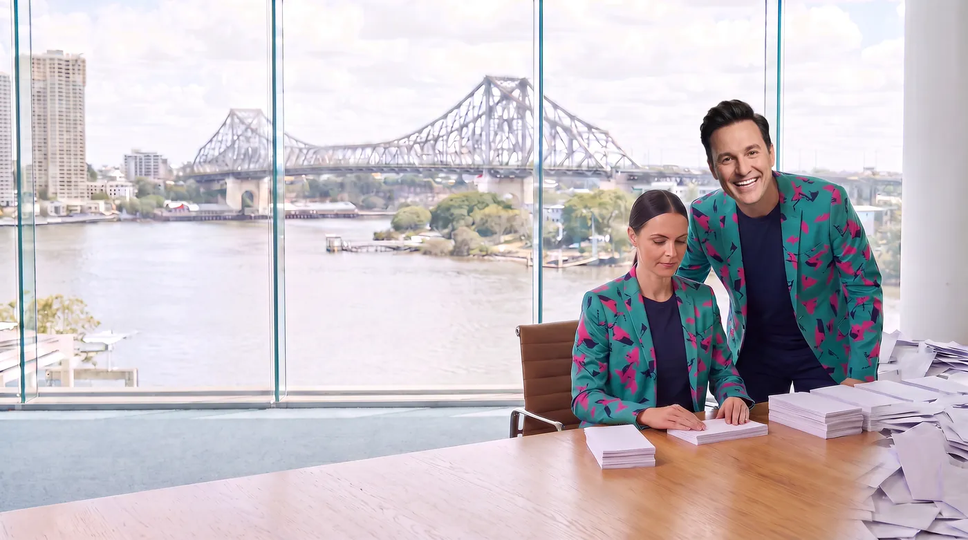 An AI staffer squares neat stacks of envelopes at a desk by the river window while Buddy (AI) leans in behind her with a grin.