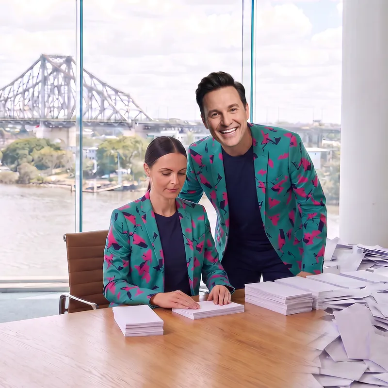 An AI staffer squares neat stacks of envelopes at a desk by the river window while Buddy (AI) leans in behind her with a grin.