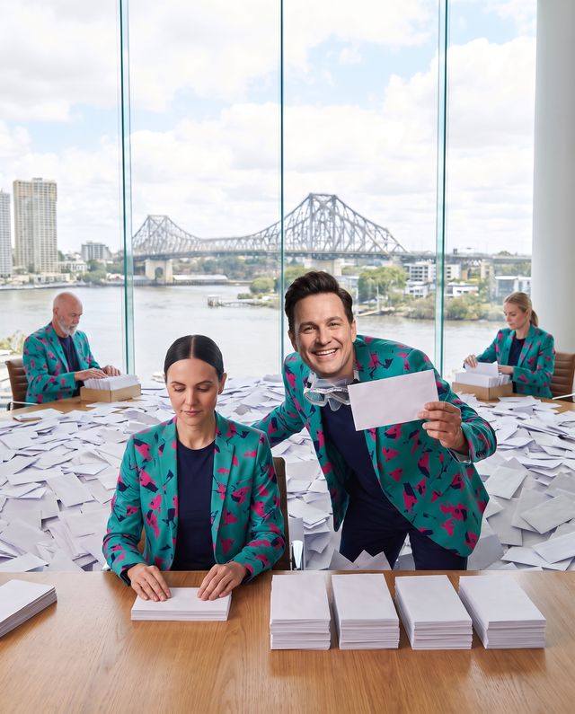An AI staffer squares a neat stack of envelopes at a desk by the river window while Buddy (AI) leans in beside her holding up a letter, the Story Bridge behind them.